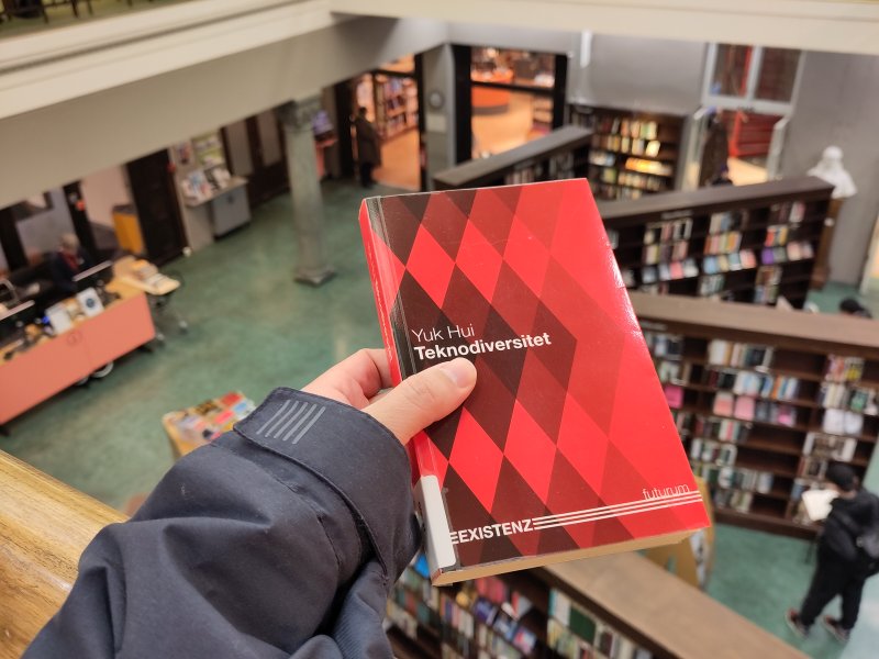 raud bok som blir holdt opp av ei hand, på eit bibliotek.